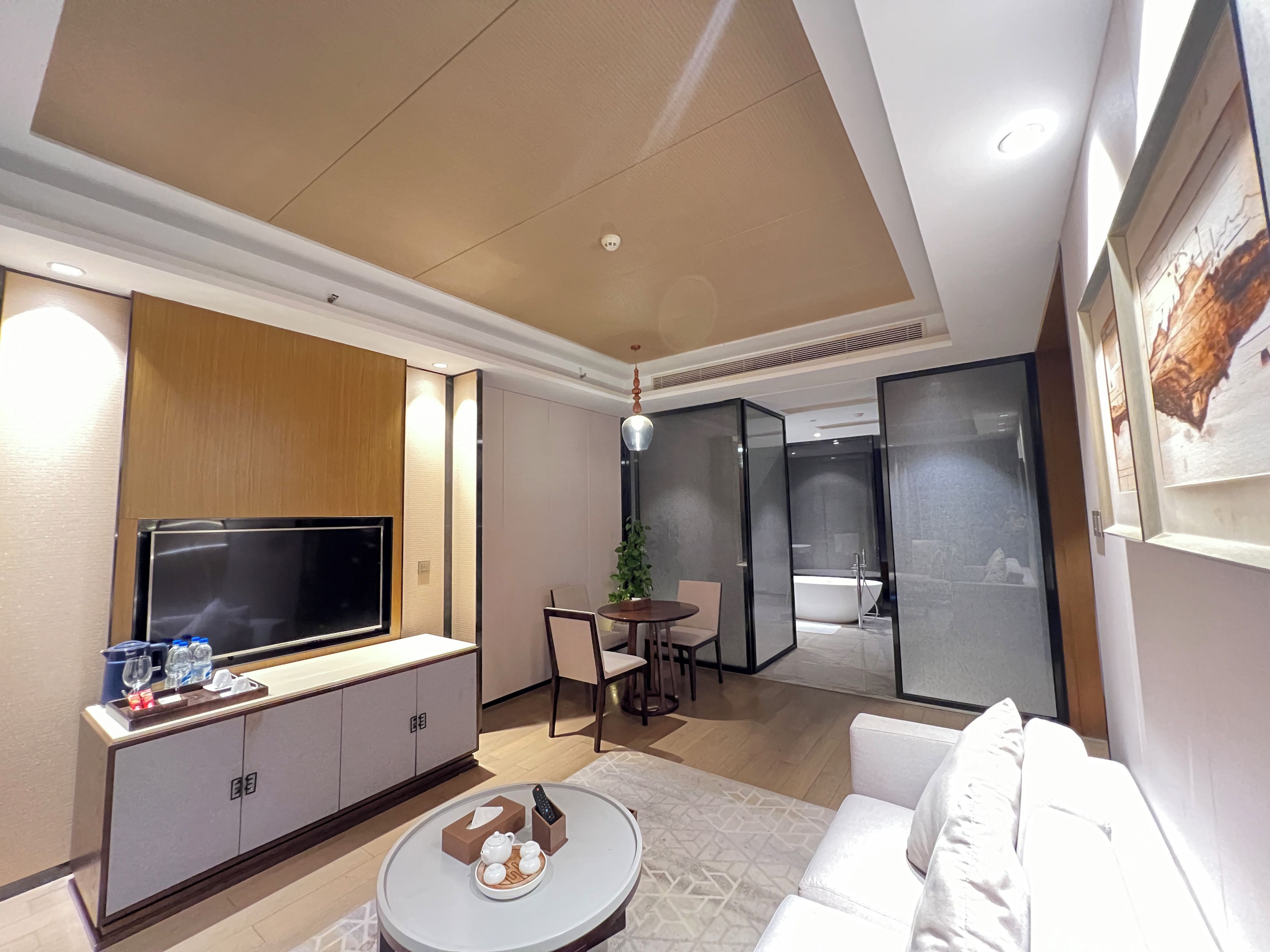 A modern hotel room with a flat-screen TV, a sofa, a dining area with a table and chairs, and a separate bathroom with a bathtub.