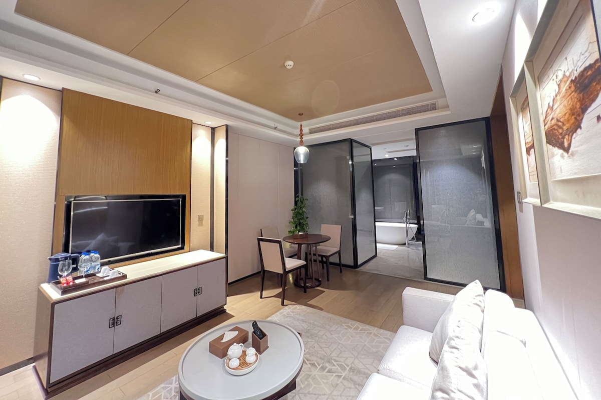 A modern hotel room with a flat-screen TV, a sofa, a dining area with a table and chairs, and a separate bathroom with a bathtub.