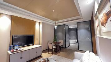A modern hotel room with a flat-screen TV, a sofa, a dining area with a table and chairs, and a separate bathroom with a bathtub.