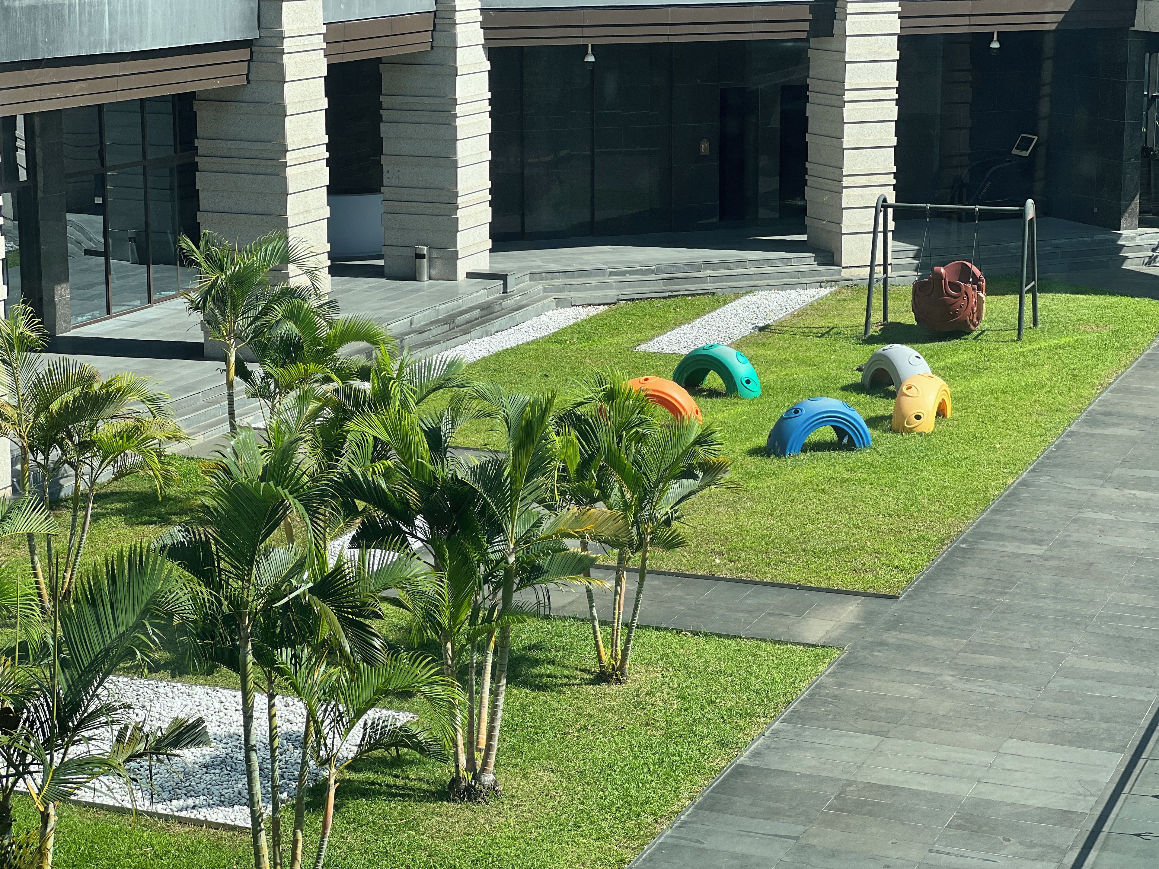 Un edificio moderno con un’area d’ingresso pavimentata, palme e colorate strutture giochi sull’erba.