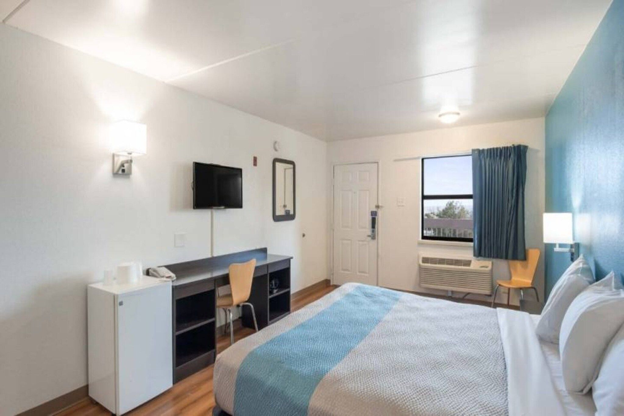 A hotel room with a bed, a desk, a chair, a television, a window with curtains, and a door.