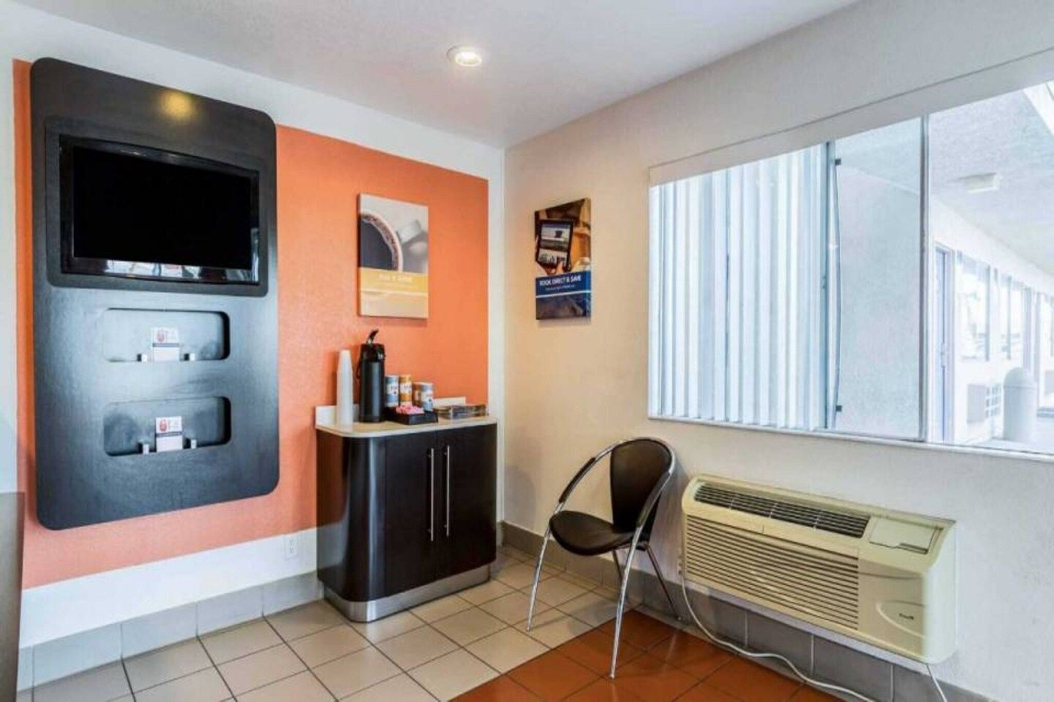 A room with a television, a small counter with a coffee machine, a chair, and a window with blinds.