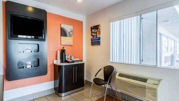 A room with a television, a small counter with a coffee machine, a chair, and a window with blinds.