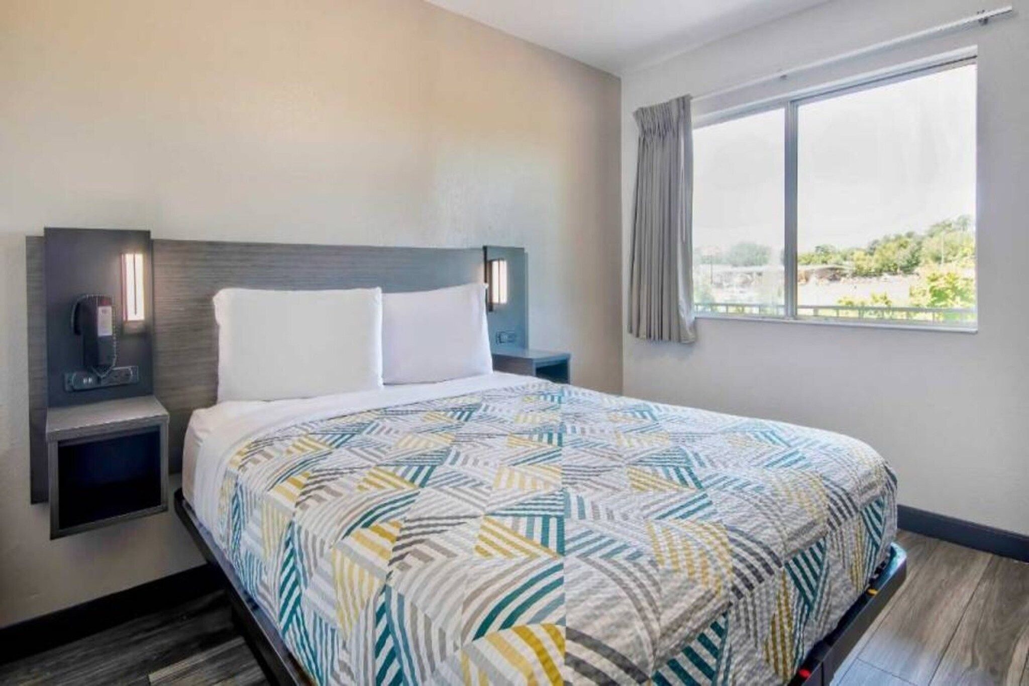 Habitación de hotel con cama, mesita de noche, lámpara de pared, ventana con cortinas y vista a zonas verdes.
