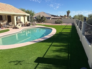 A swimming pool with artificial grass, a covered patio with chairs, and a view of a residential area with houses and trees. - Charming 3 Bedroom 2 Bath Home in Fountain Hills with Pool and Mountain Views! (Fountain Hills)
