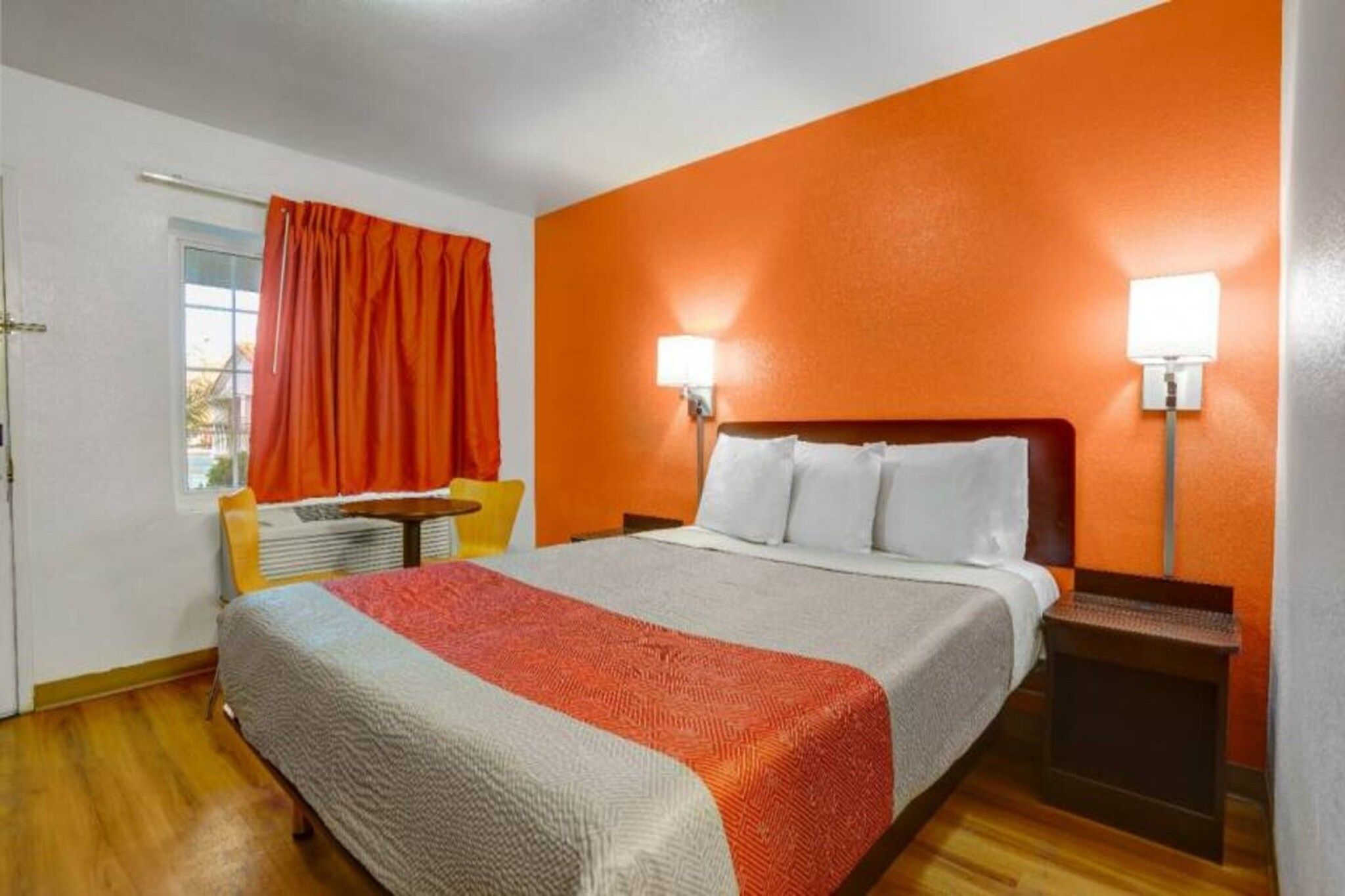 A hotel room with a bed, a nightstand, a chair, a window with curtains, and a wall lamp.
