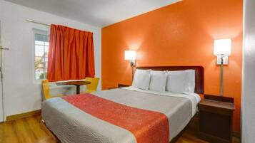 A hotel room with a bed, a nightstand, a chair, a window with curtains, and a wall lamp.