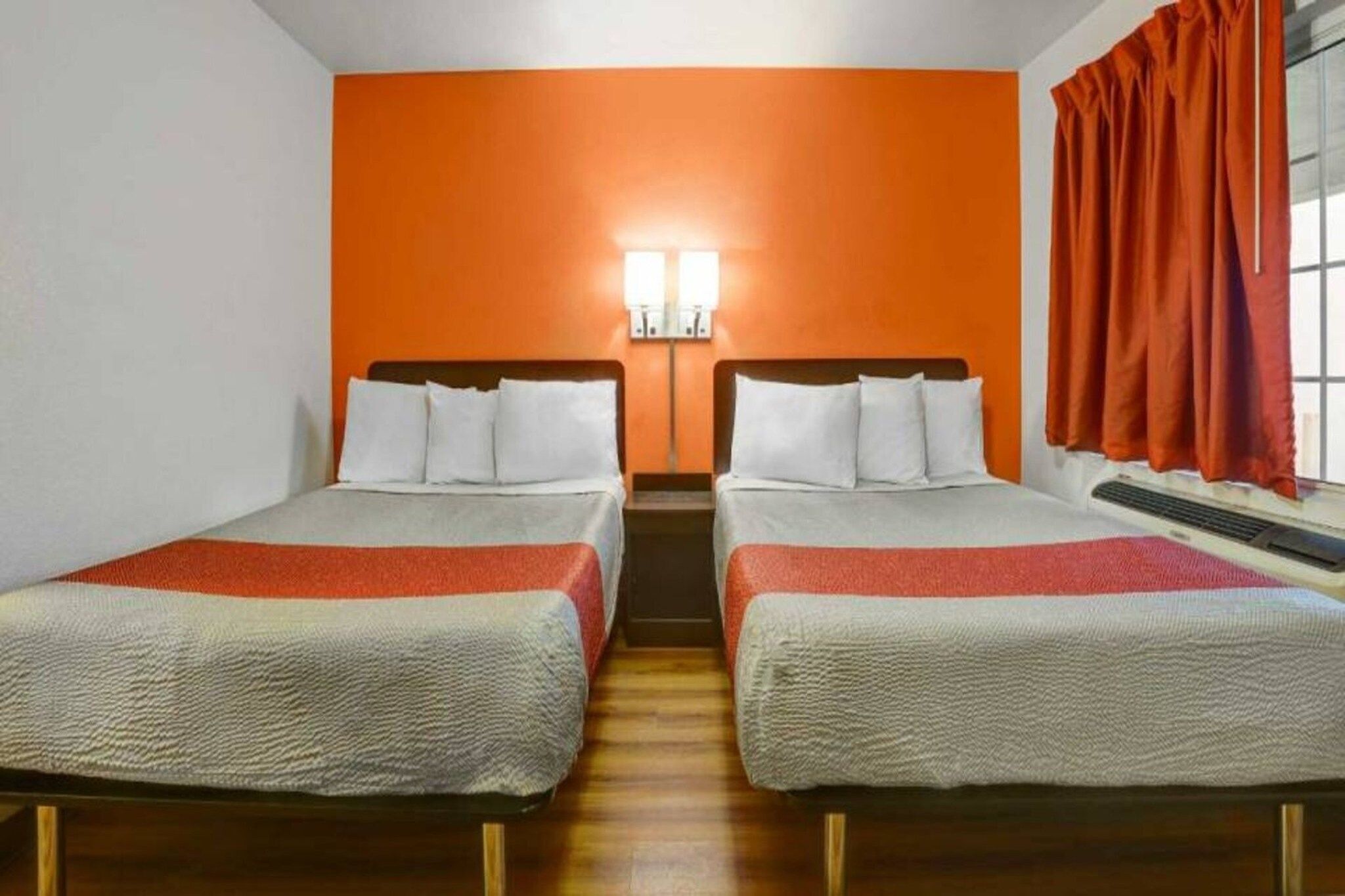 A hotel room with two beds, an orange accent wall, and a window with curtains.
