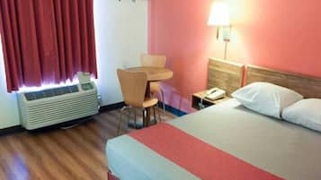 A hotel room with a bed, a desk, a chair, an air conditioner, a lamp, and a telephone.