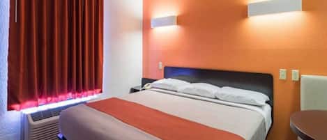 A hotel room with a bed, a bedside table, a chair, a heater, and a window with curtains.