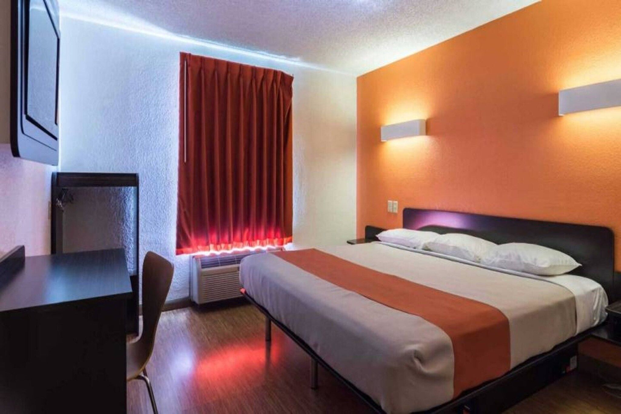 Quarto de hotel com cama, mesas de cabeceira, escrivaninha, cadeira, janela com cortinas e luminária na parede.