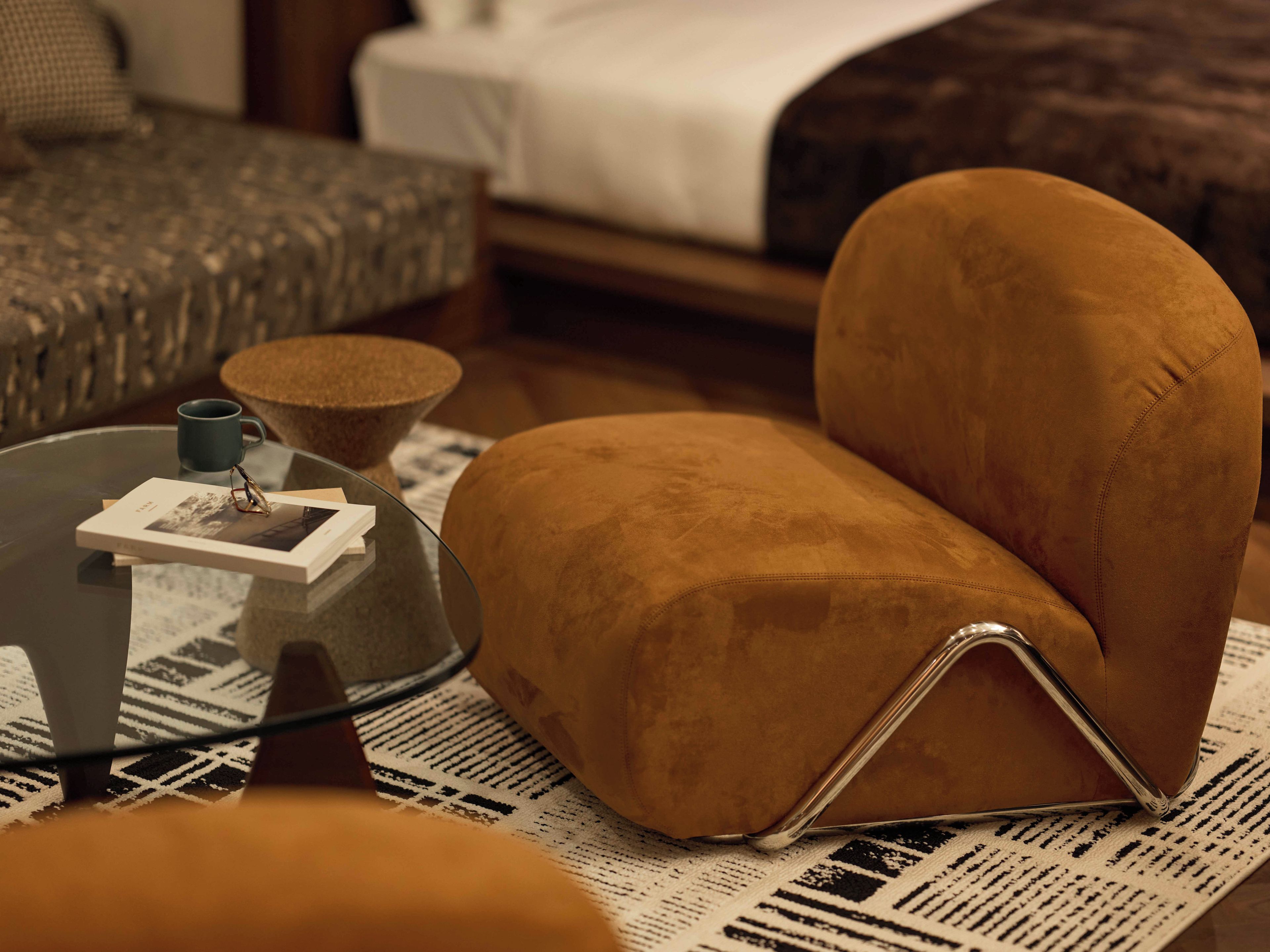 A cozy room with a brown armchair, a glass coffee table with a book and glasses, and a patterned rug.