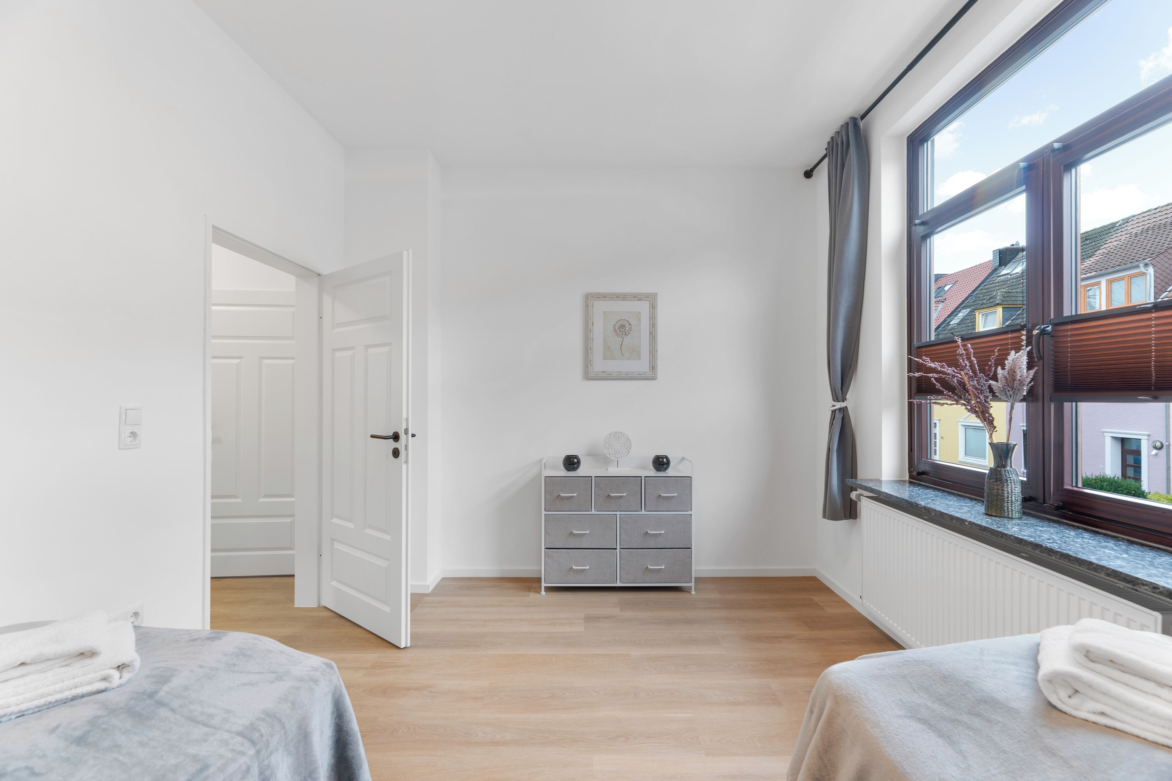 A bedroom with two beds, a dresser, a window with curtains, and a door leading to another room.