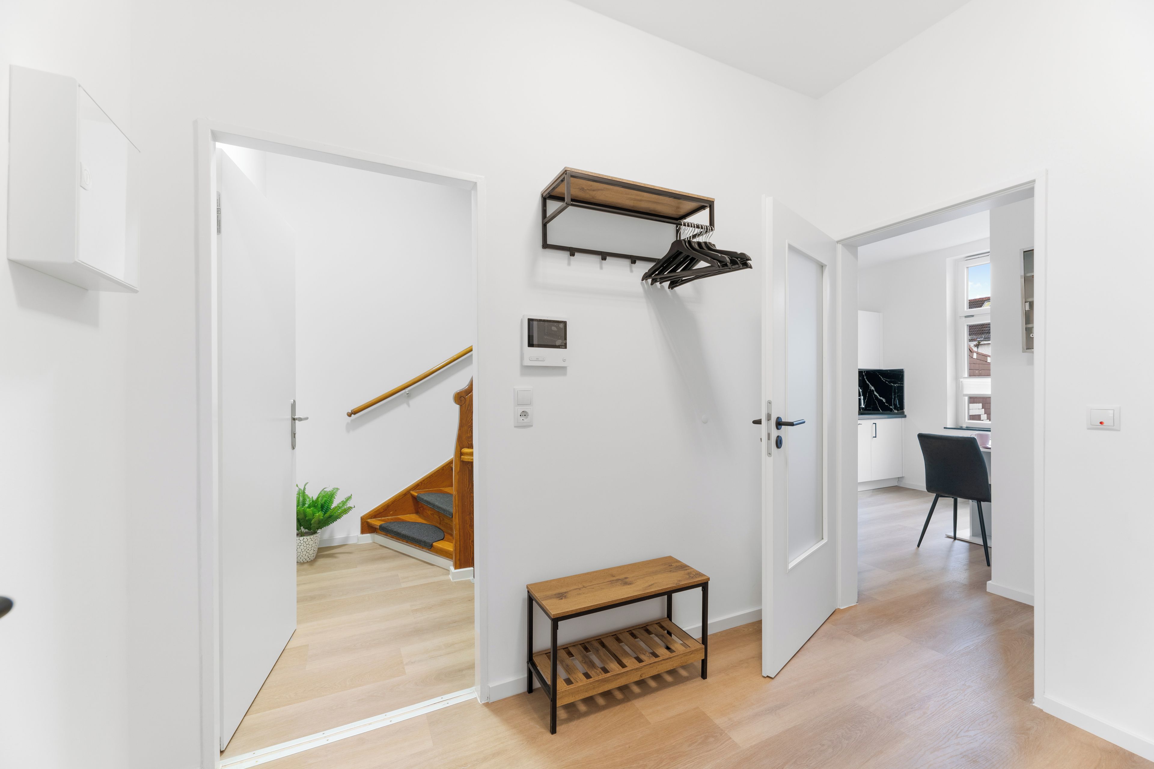 A modern interior with a wooden staircase, a coat rack, a small bench, and a dining area with a black chair.