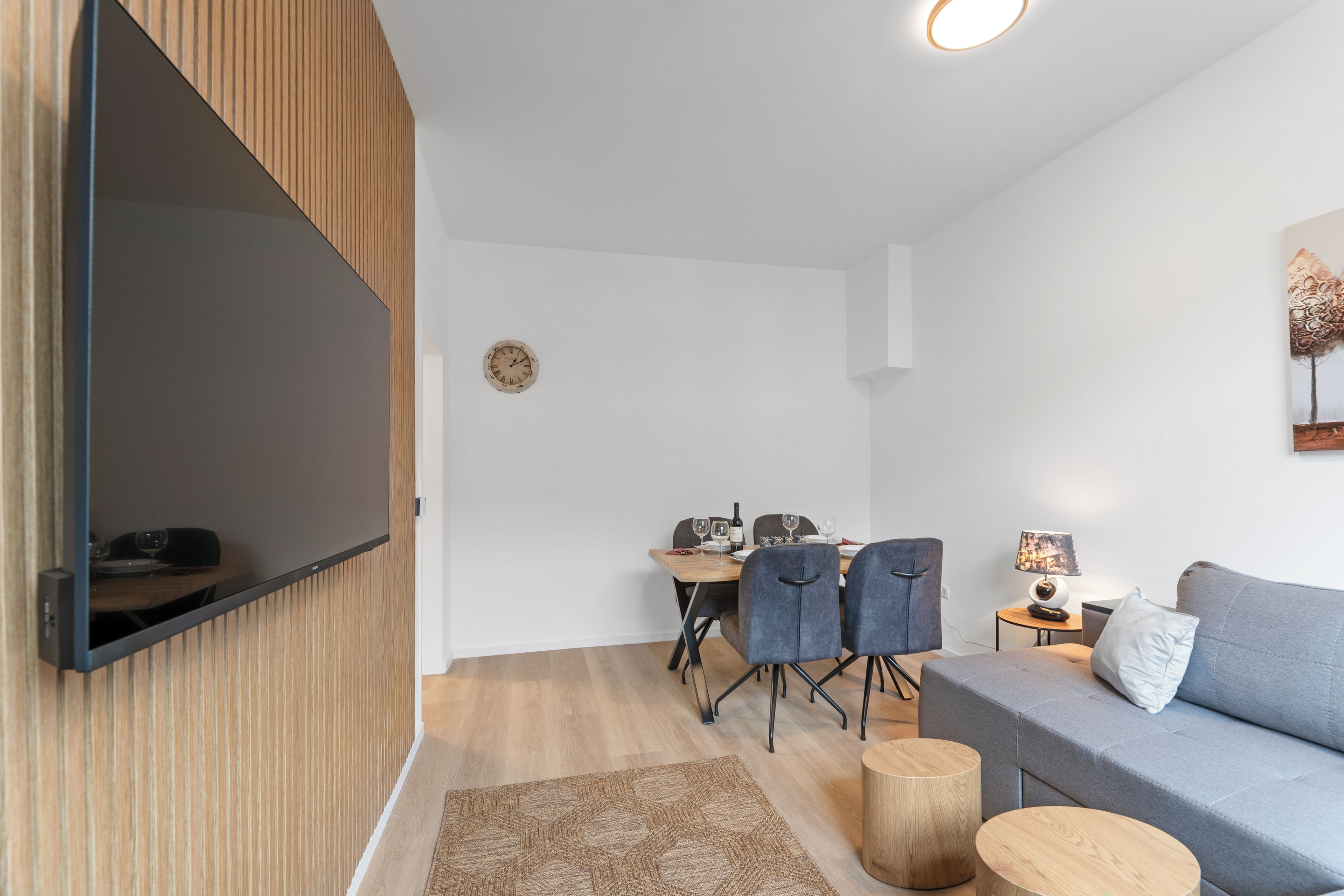A modern living room with a flat-screen TV mounted on the wall, a dining area with a table and chairs, and a grey sofa with a side table.