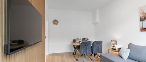 A modern living room with a flat-screen TV mounted on the wall, a dining area with a table and chairs, and a grey sofa with a side table.
