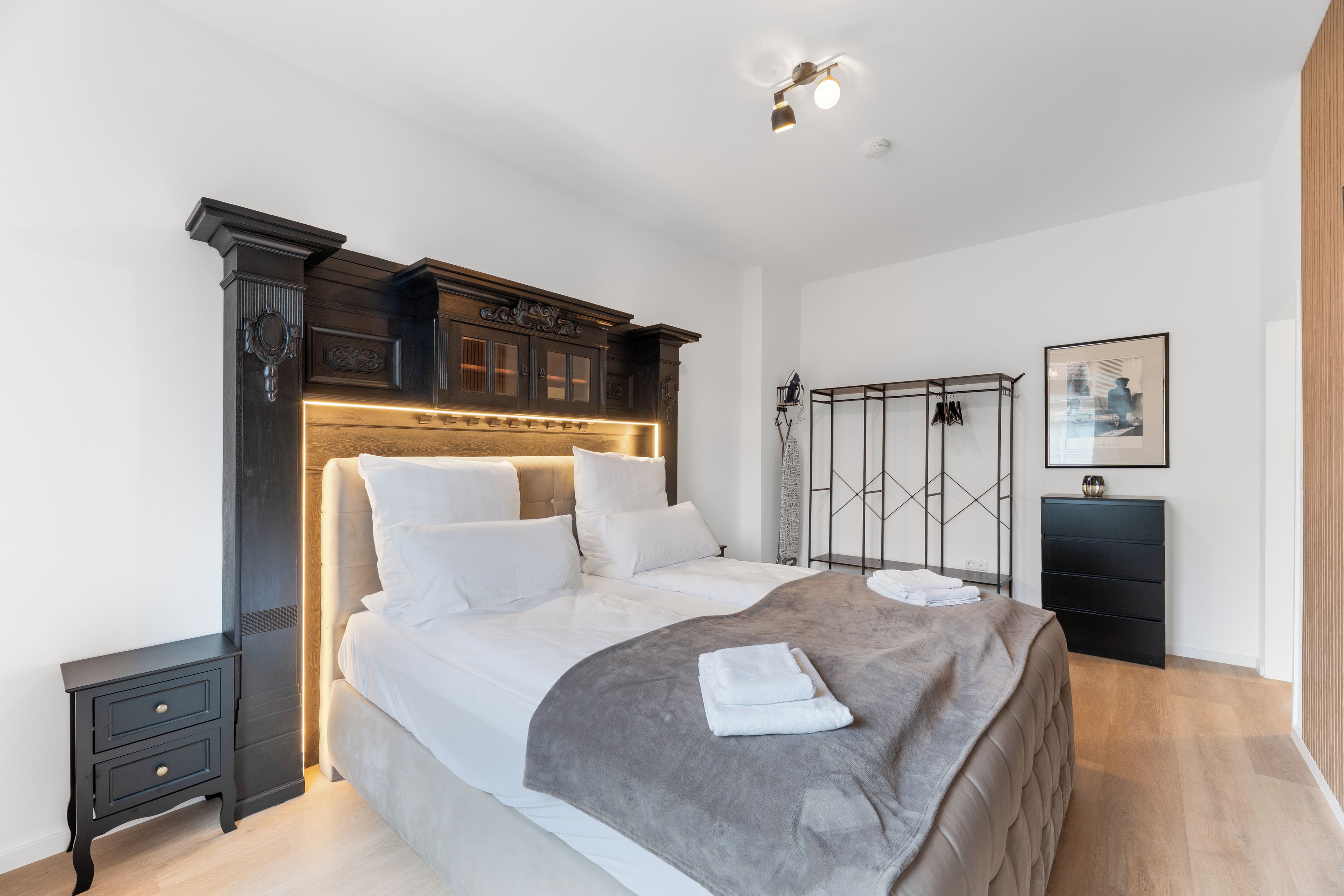 Ein modernes Schlafzimmer mit einem großen Bett, einem hölzernen Kopfteil, einem Nachttisch, einem Kleiderschrank und einem gerahmten Bild an der Wand.