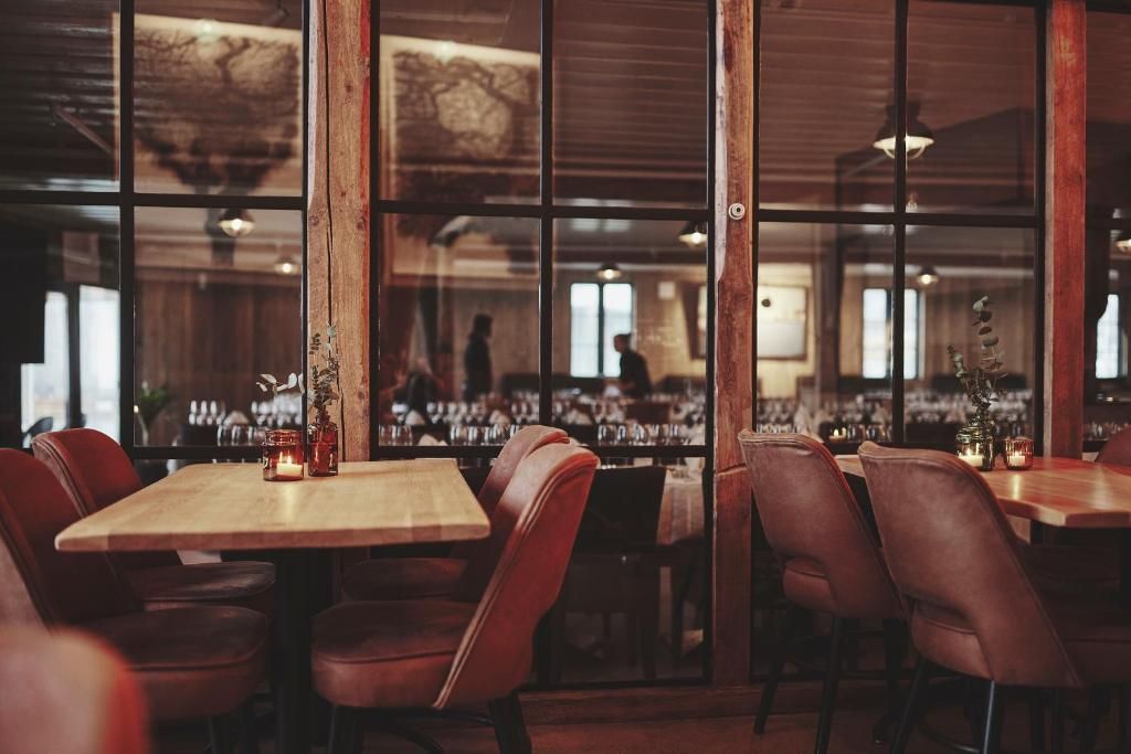 Et restaurantinteriør med træborde og stole, store vinduer og et barområde i baggrunden.