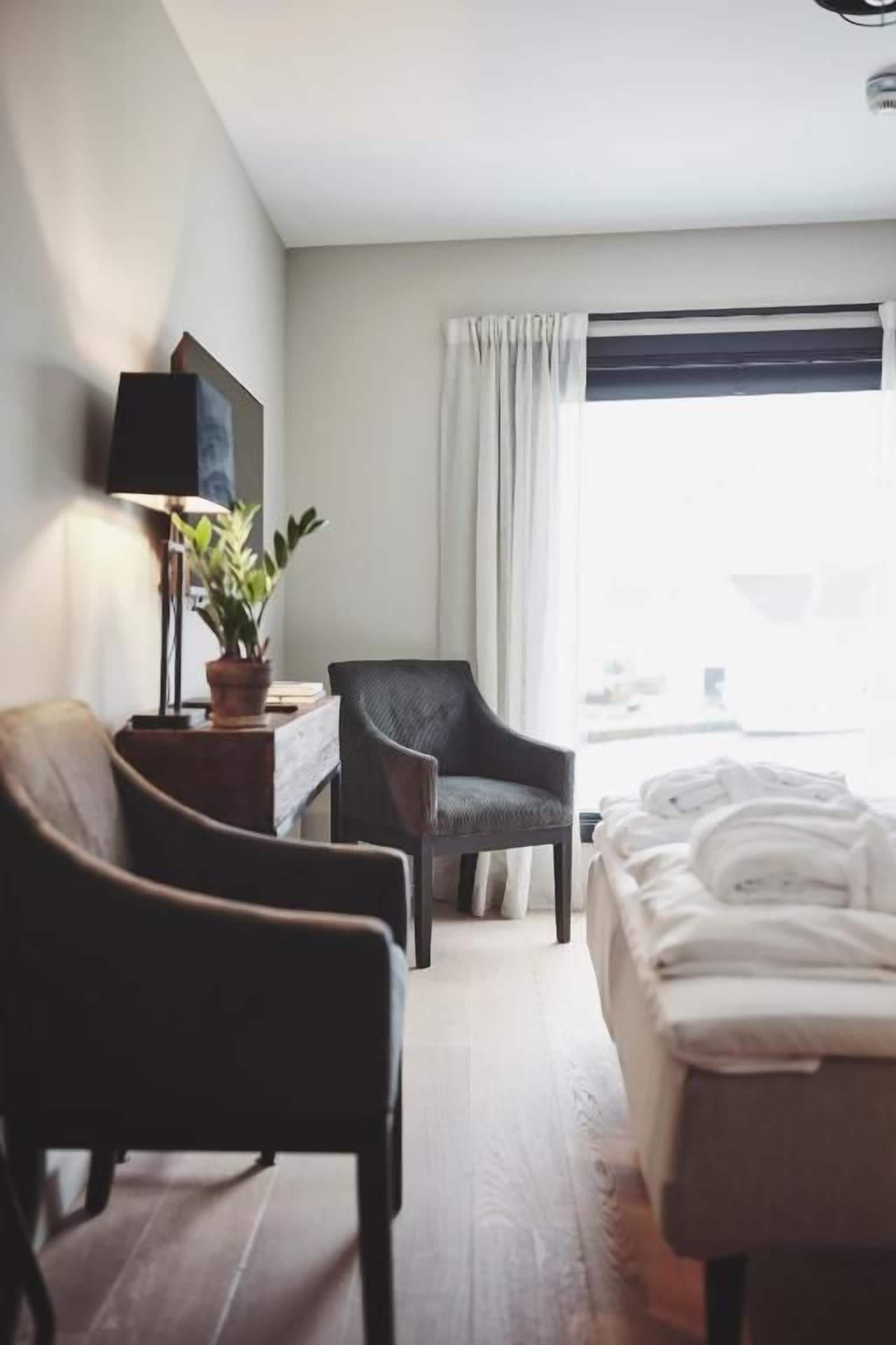 A hotel room with a bed, a chair, a desk with a lamp, and a window with curtains.