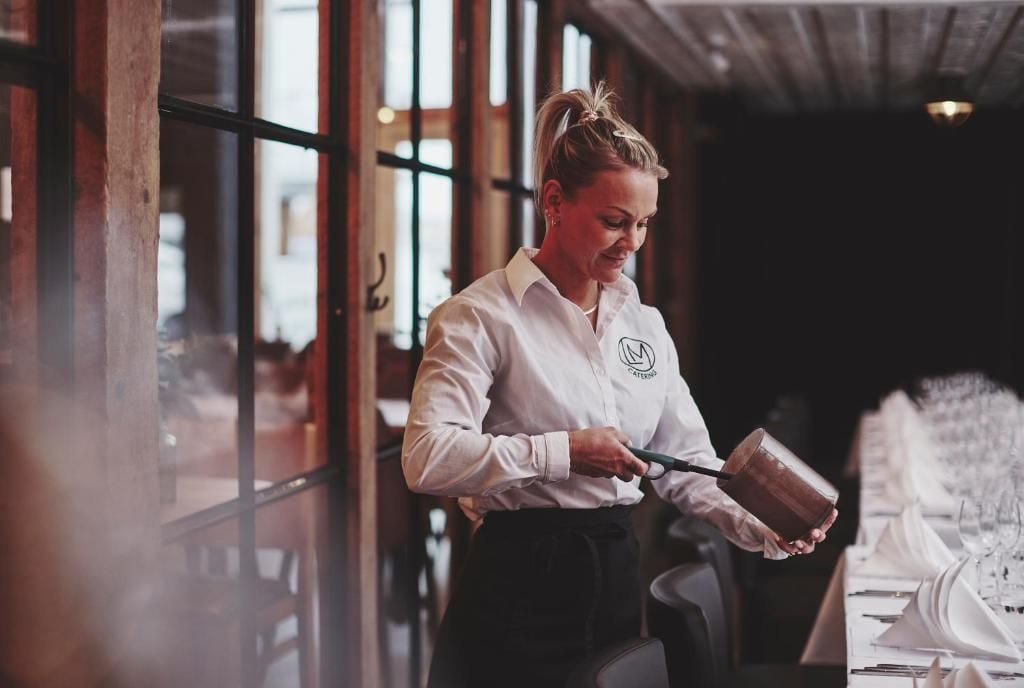 A person in a white shirt with a logo, holding a container, standing in a room with tables set for dining.