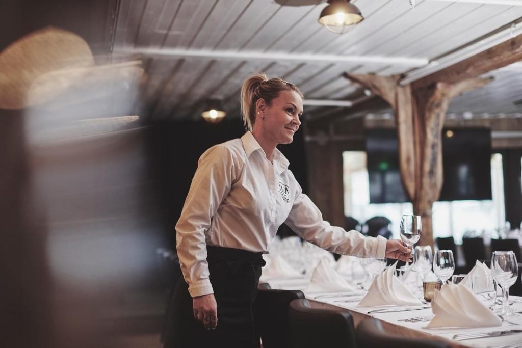 A person setting a table with white tablecloths and silverware in a restaurant with wooden beams and pendant lighting.