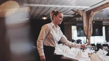 A person setting a table with white tablecloths and silverware in a restaurant with wooden beams and pendant lighting.