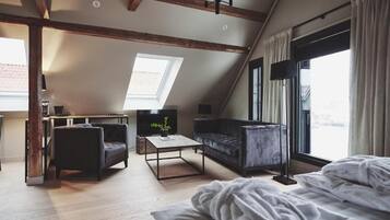 A loft-style bedroom with a sloped ceiling, a large skylight, a bed with white bedding, a dark sofa, a coffee table, and a TV.