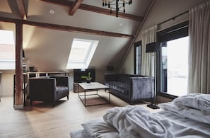 Une chambre de style « loft » avec un plafond en pente, une grande lucarne, un lit avec une literie blanche, un canapé foncé, une table basse et un téléviseur.
