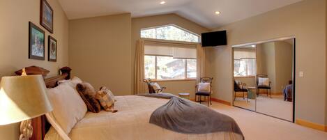 A bedroom with a large bed, a bedside table with a lamp, a chair, a window with blinds, and a mirror on the wall.