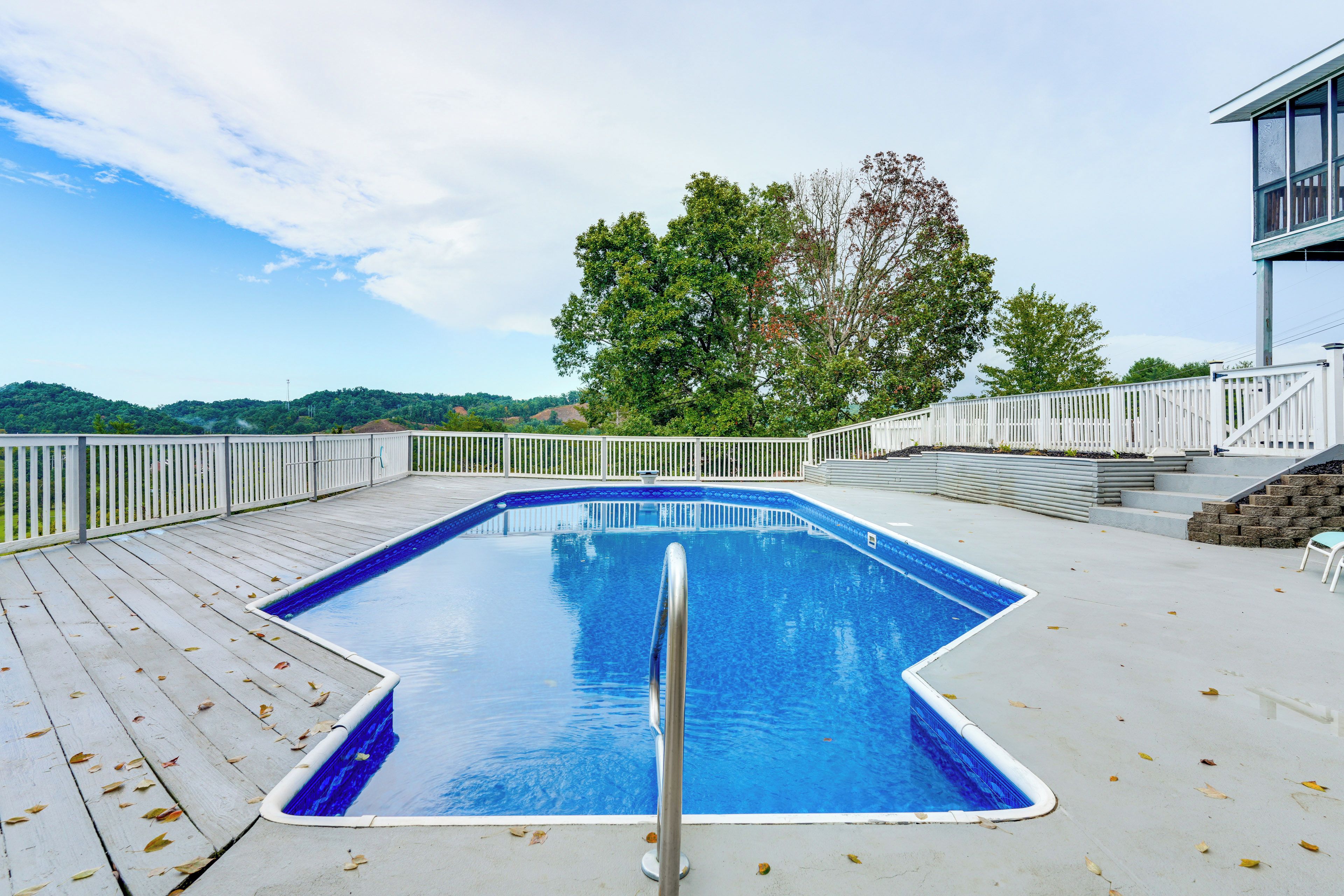 Una piscina rectangular con una escalera metálica, rodeada por un patio blanco con barandilla blanca, y vistas a una colina con árboles.