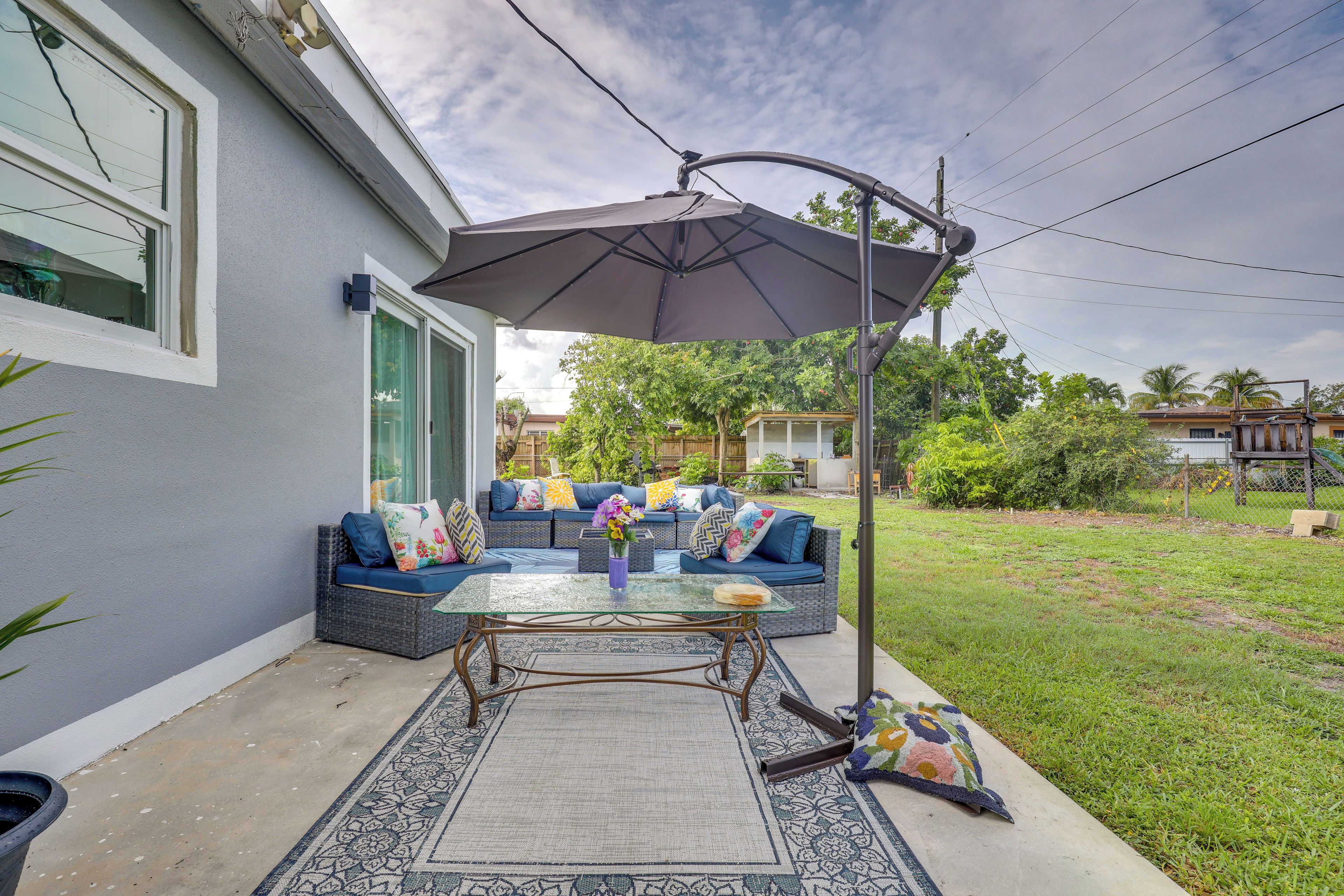 An outdoor patio area with a glass-top table, a gray sofa with colorful cushions, and a large umbrella.