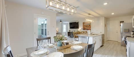 A dining area with a wooden table set for four, a kitchen in the background, and a ceiling light fixture.