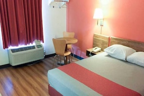 A hotel room with a bed, a desk, a chair, an air conditioner, a lamp, and a telephone.