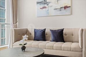 A beige sofa with two dark cushions, a small round table with a potted plant, and a window with curtains. - Niro by Kozystay - Kuningan (South Jakarta)