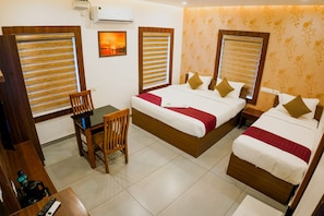 A hotel room with a bed, a desk, and a chair. There is a window with blinds, a wall-mounted air conditioner, and a framed picture on the wall.
