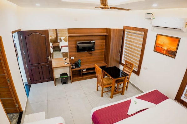 A compact hotel room with a bed, a desk, a chair, a TV, and a window with blinds.