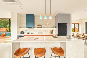 A modern kitchen with a central island, bar stools, pendant lighting, and a view of the outdoors. - The RED Signature 5br sunset (Đà Nẵng)