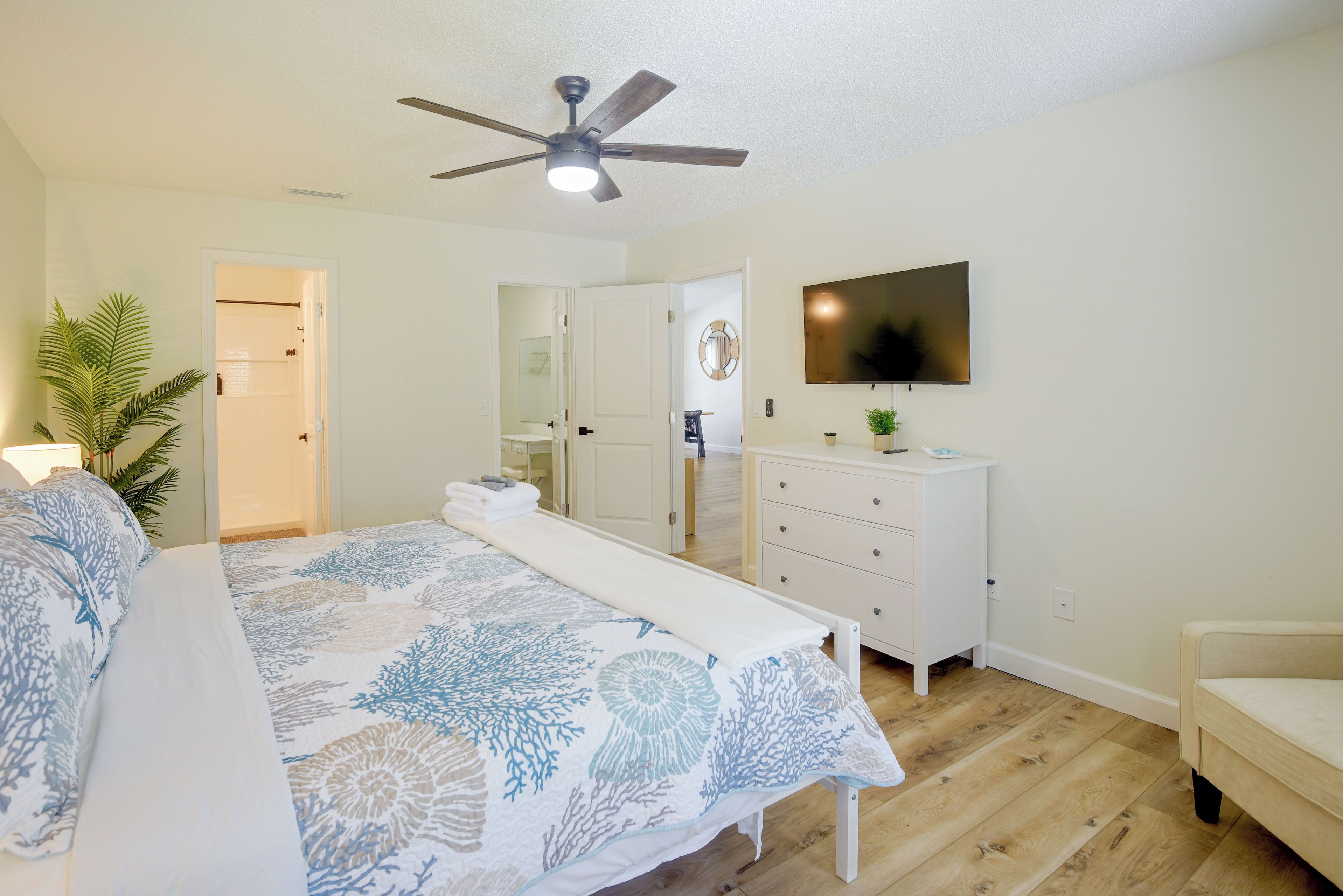 A bedroom with a bed, a ceiling fan, a TV, a dresser, and a plant.