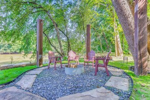 An outdoor seating area with Adirondack chairs around a fire pit, surrounded by a stone border and trees.