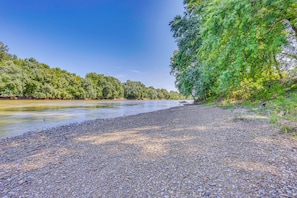 A pebbled riverbank with dense greenery on one side and a clear blue sky.
