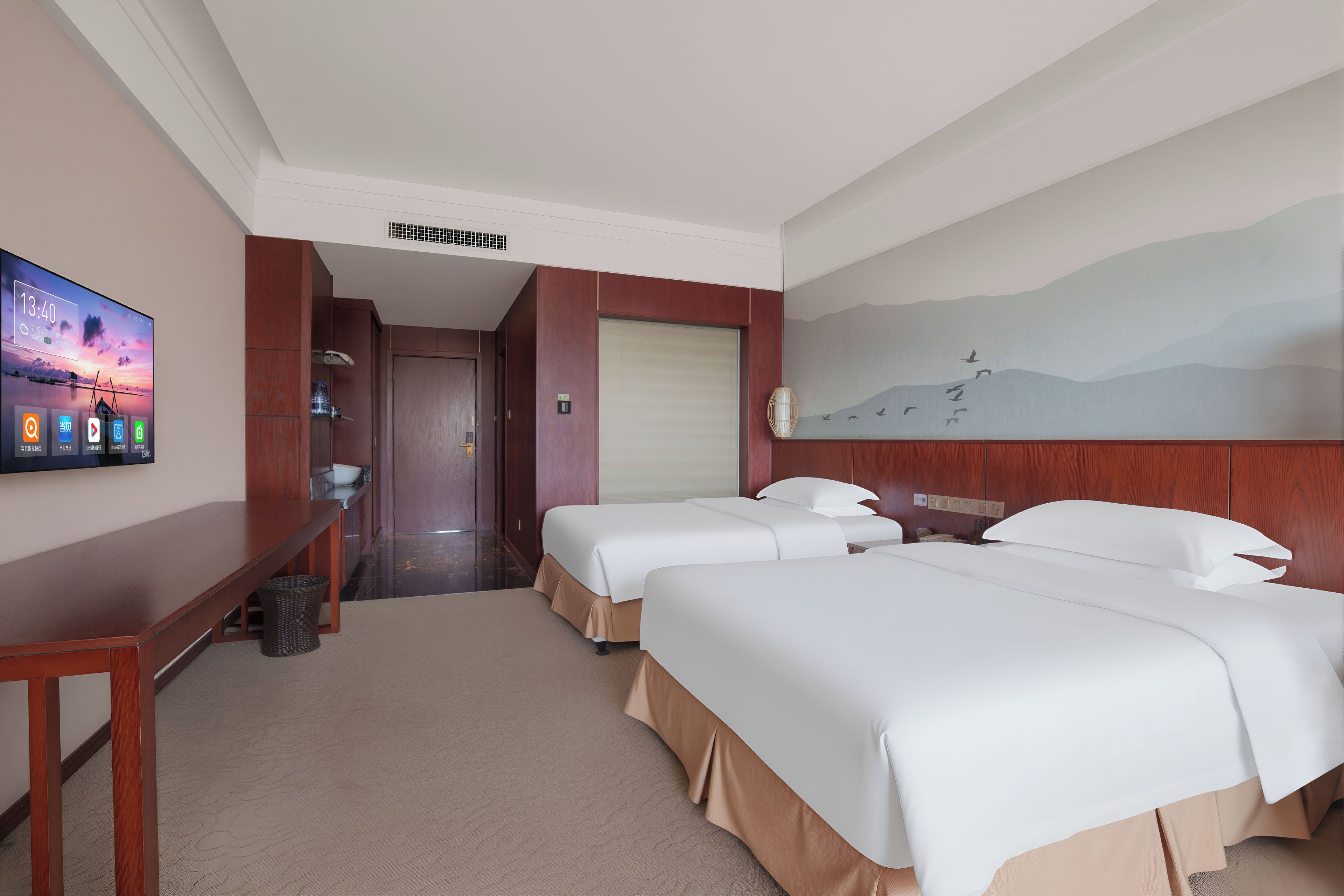 Een hotelkamer met twee bedden, een bureau, een televisietoestel waarop de tijd staat en een schilderij aan de muur.