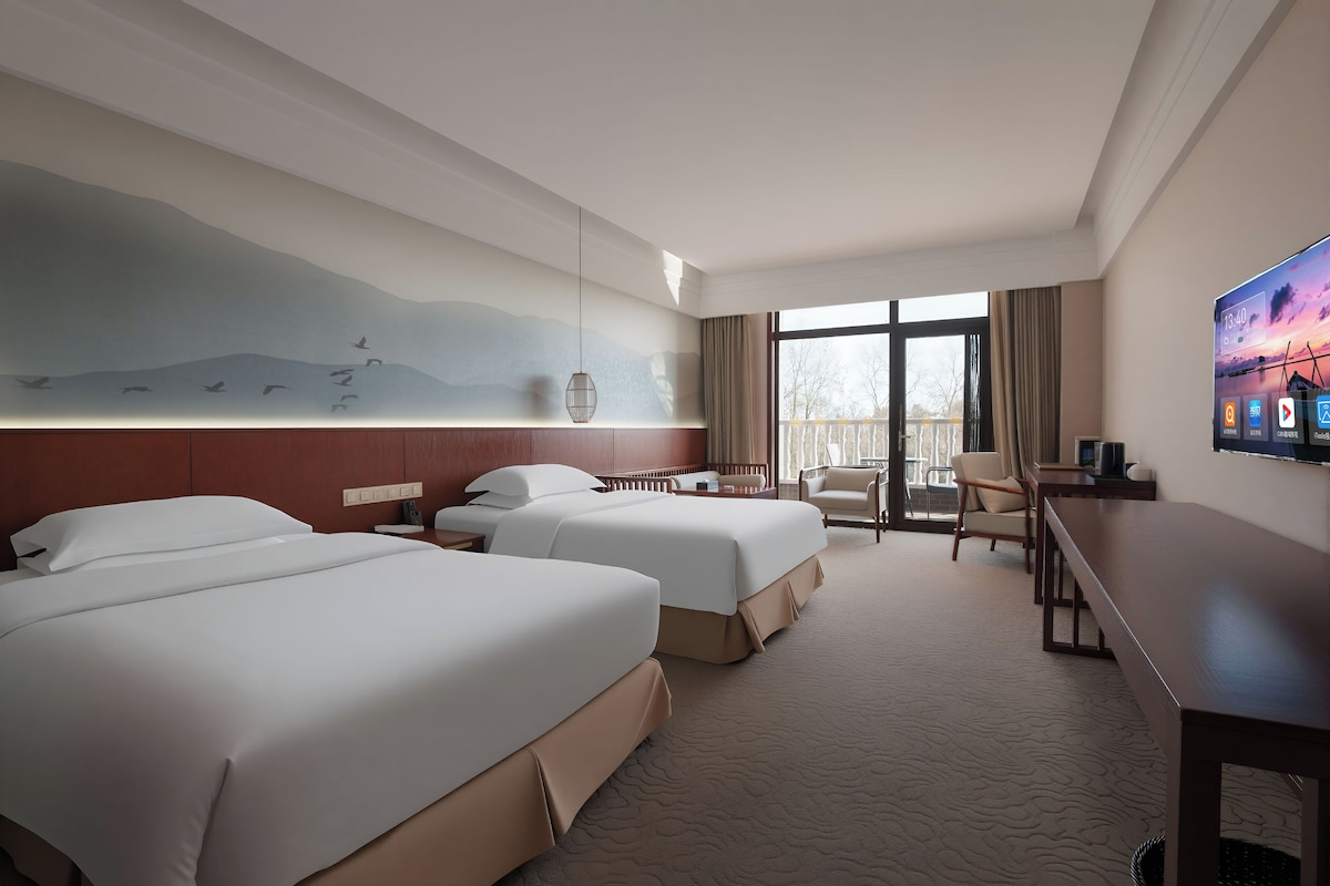 A hotel room with two beds, a desk, a chair, a TV, and a balcony with a view.