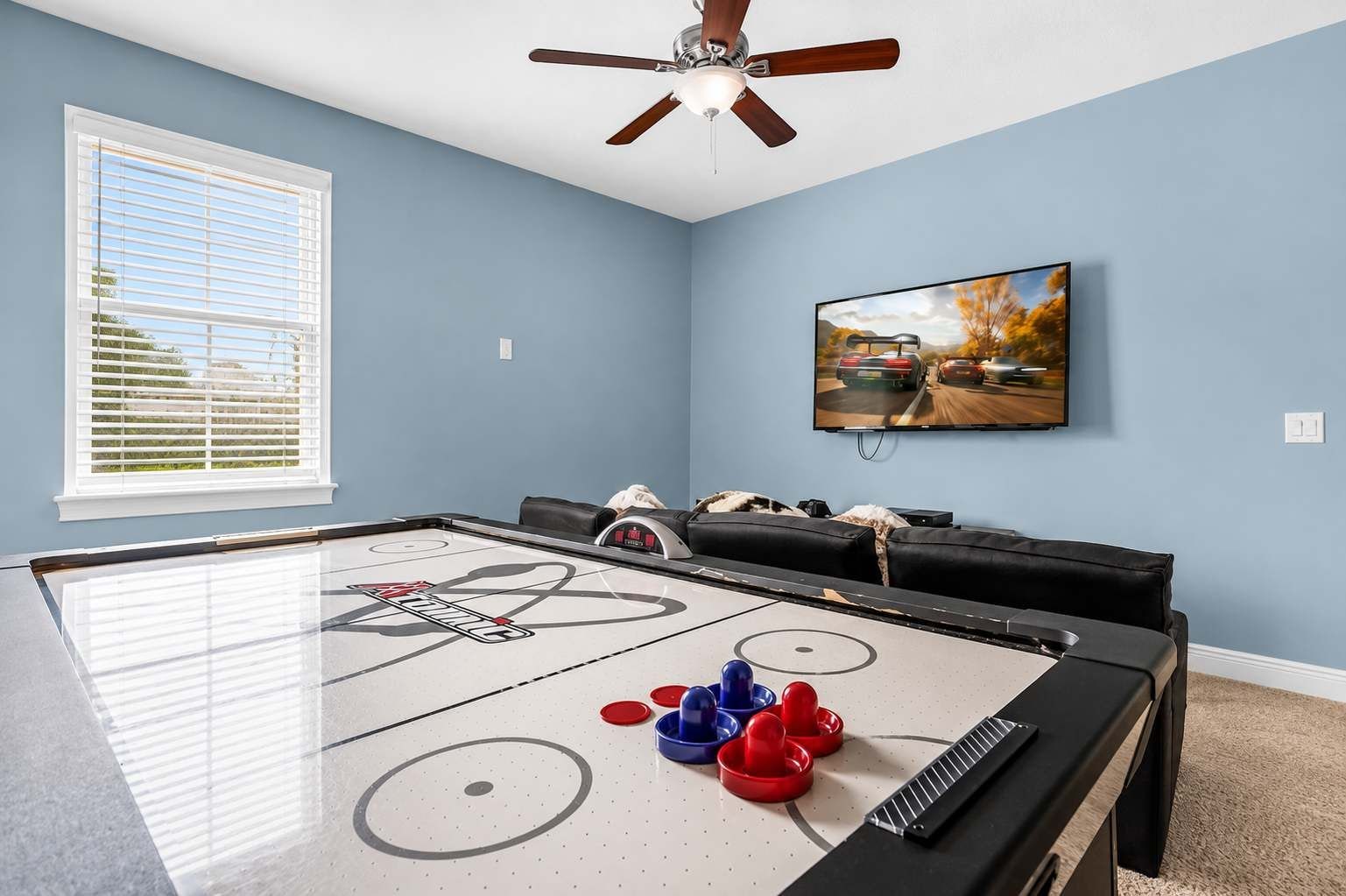 A room with a shuffleboard table, a ceiling fan, a television, and a window with blinds.