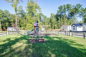 A playground with a slide and climbing structures, surrounded by a fence and grassy area. - Communal Fire Pit & Dock! Tiny Home By Lake Murray (Gilbert)