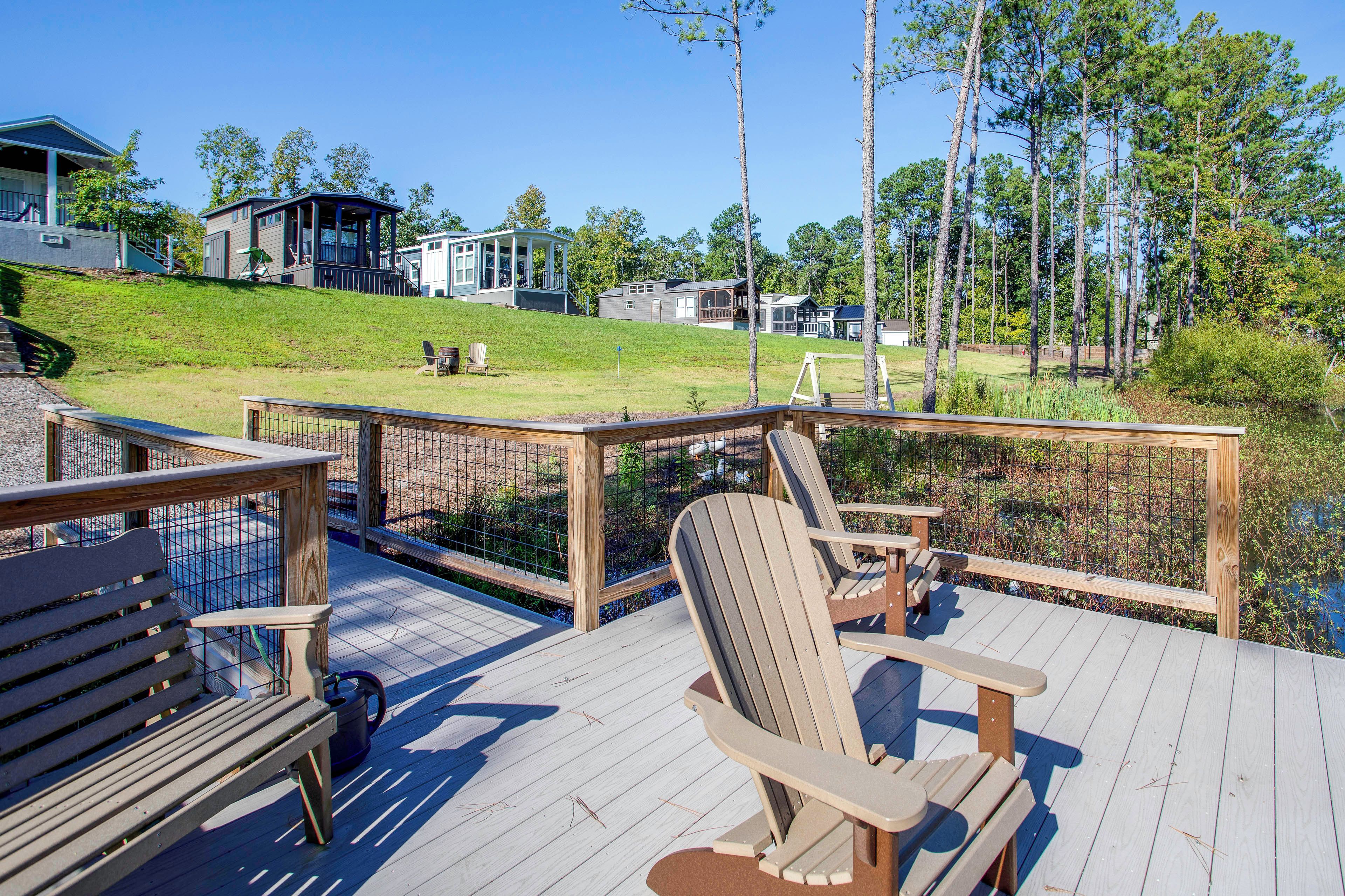 Um deck de madeira com cadeiras Adirondack e um banco, com vista para uma área gramada com casas e árvores.