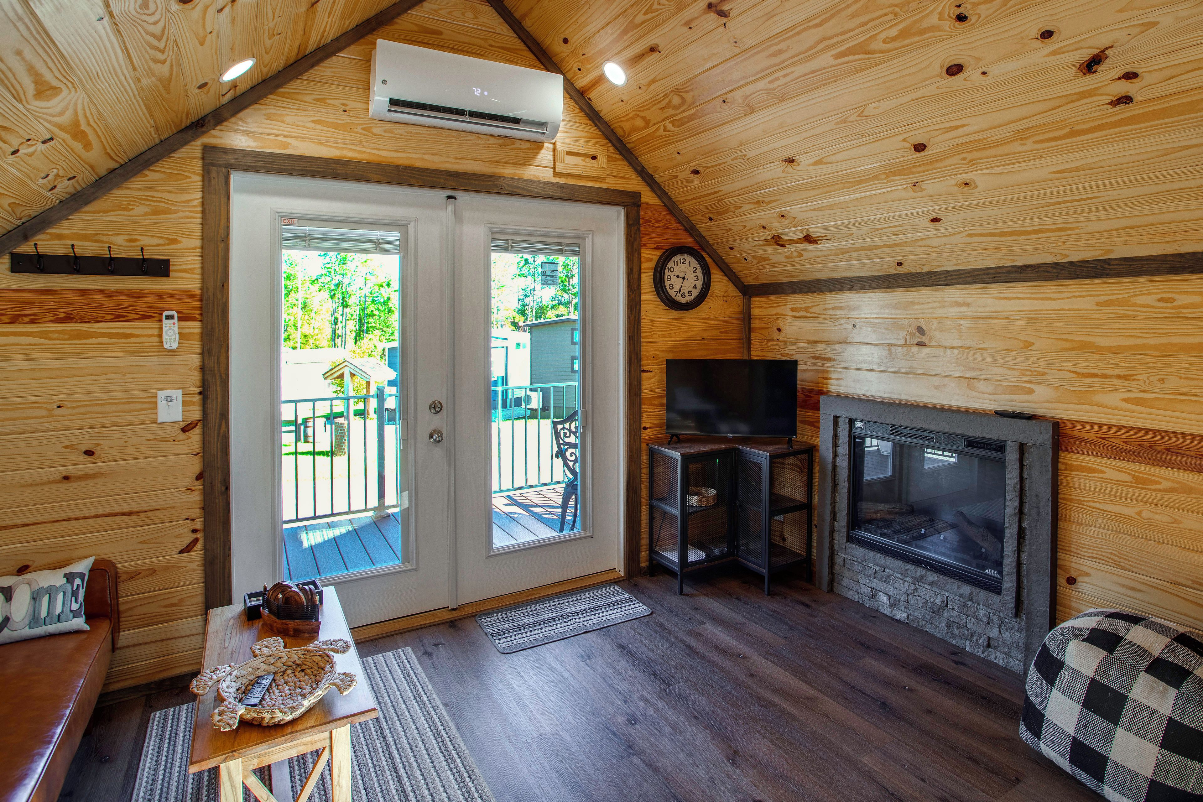 Ein gemütliches Blockhaus-Interieur mit Holzbalkendecke, Kamin, Fernseher und einer Tür zu einer Außenterrasse.
