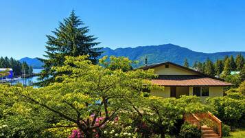 A house with a wooden staircase leading to a porch, surrounded by lush greenery and a mountain backdrop.