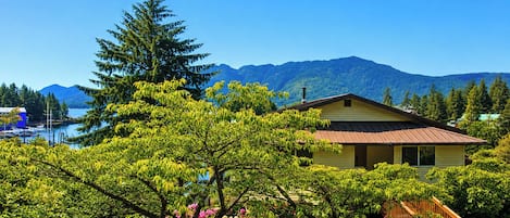 A house with a wooden staircase leading to a porch, surrounded by lush greenery and a mountain backdrop.
