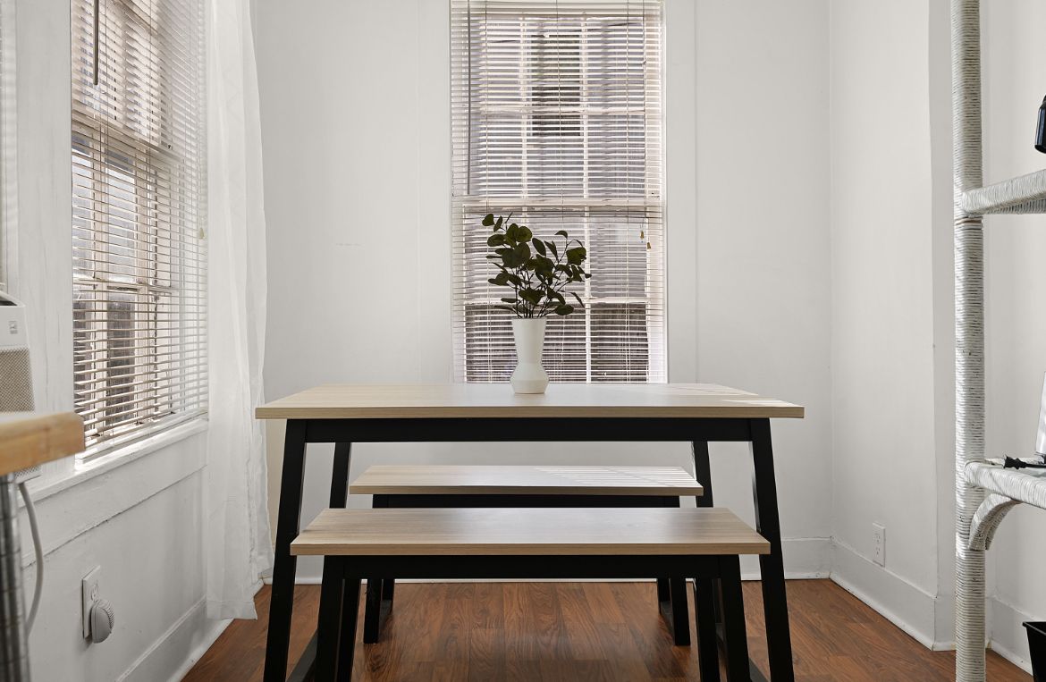 Una habitación con pared blanca, una ventana con persianas, un florero blanco con una planta, un banco de madera y un radiador blanco.