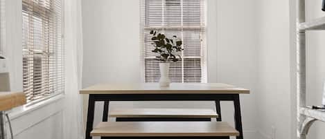 Una habitación con pared blanca, una ventana con persianas, un florero blanco con una planta, un banco de madera y un radiador blanco.